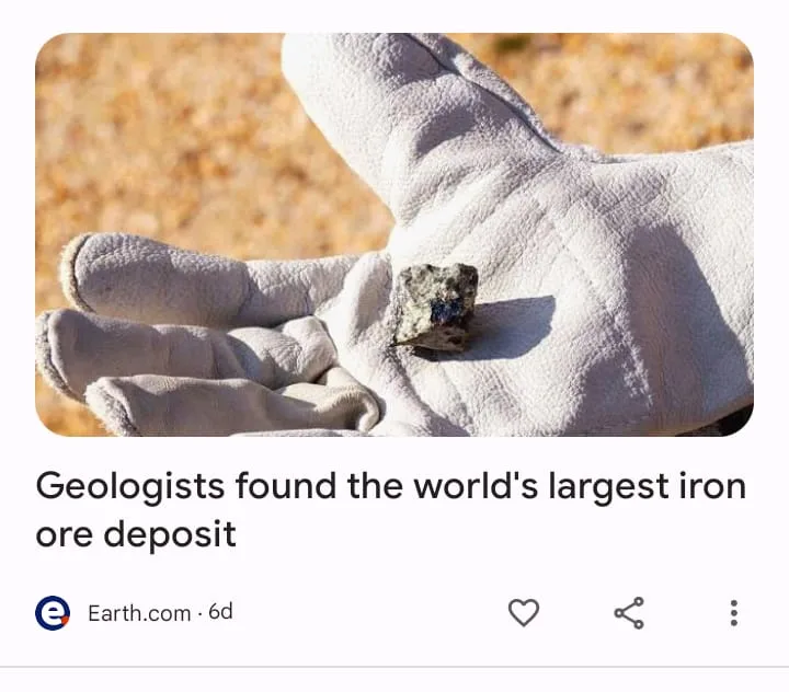 A close-up photo shows a gloved hand holding a small, rough, dark-colored rock against a blurred sandy background. The…