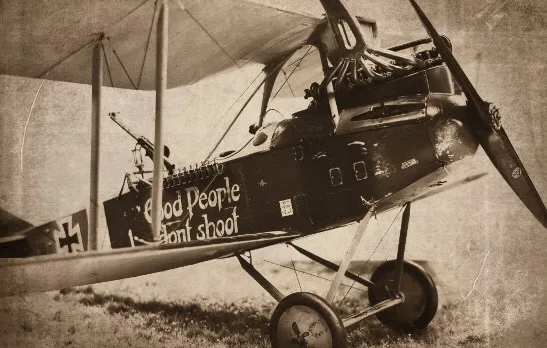 A sepia-toned historical photograph of a World War I-era German biplane, featuring a black cross insignia on its wing and…