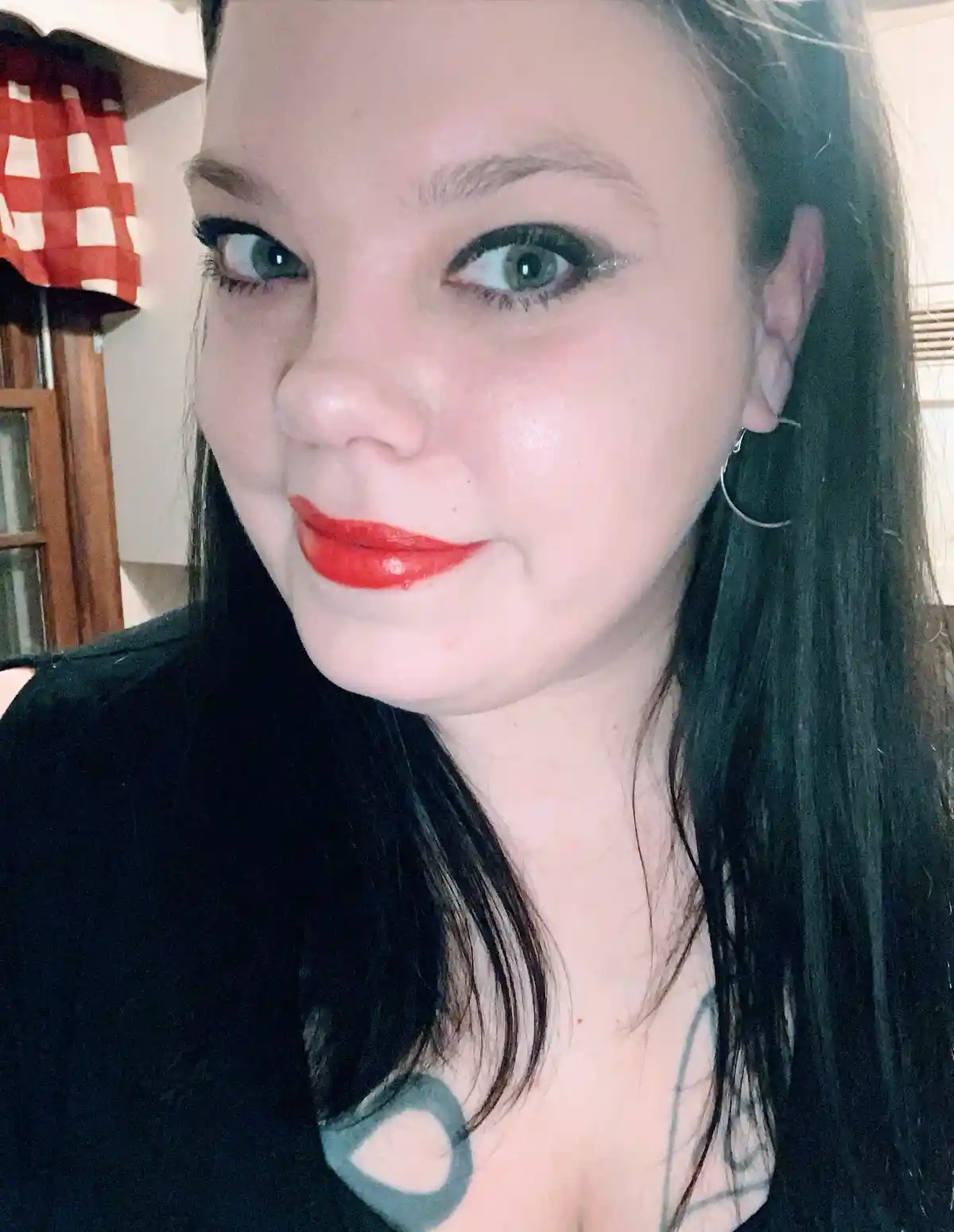A close-up selfie of a woman with dark hair, wearing bold red lipstick and dramatic dark eyeliner, looking directly at the…