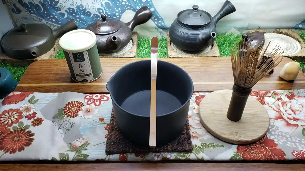 A still life of traditional Japanese tea ceremony equipment including a black bowl with a wooden spoon, a bamboo whisk, a…