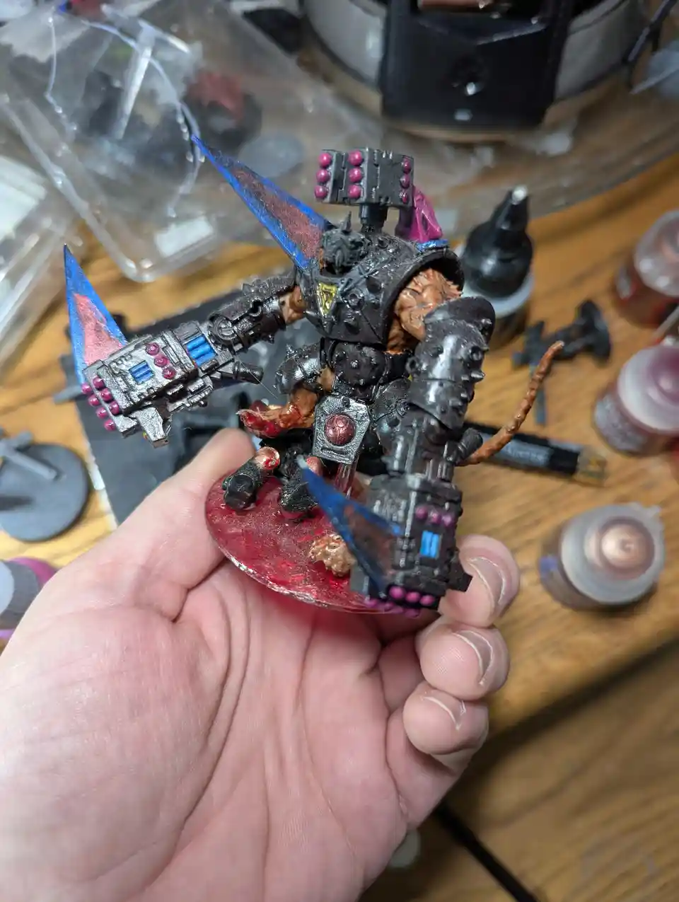 A hand holds a heavily painted, grimdark-style miniature figure with black armor, blue and pink highlights, and a red base…