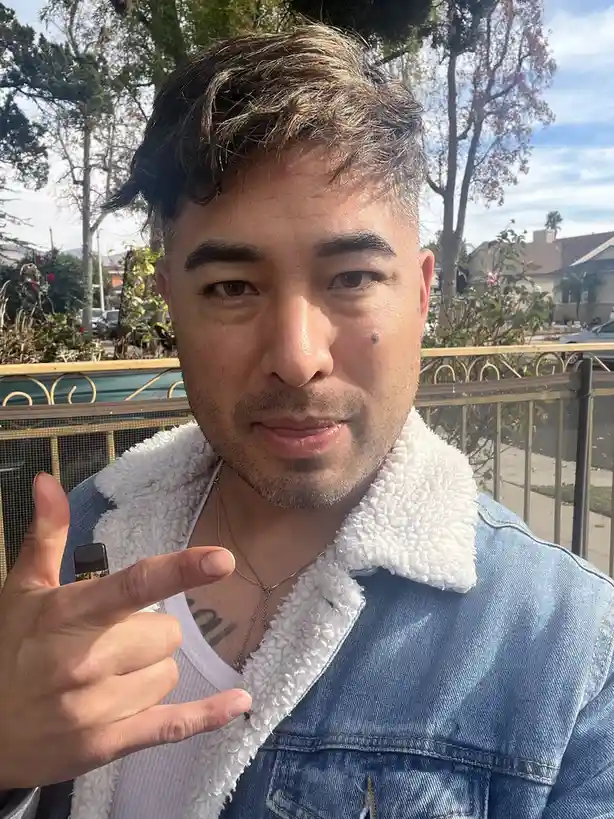 A man with styled hair and a denim jacket with a white fleece collar poses outdoors, holding a vape pen in his hand while…