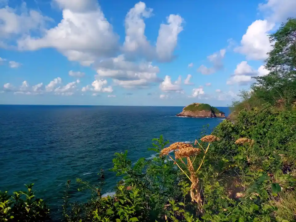 A scenic coastal view shows a calm blue ocean under a partly cloudy sky, with a small rocky island in the distance and green…