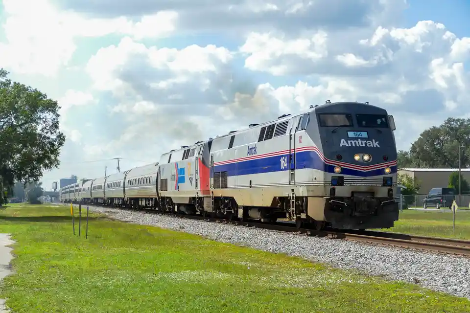An Amtrak passenger train, numbered 164, travels along a track through a grassy area under a partly cloudy sky. The…