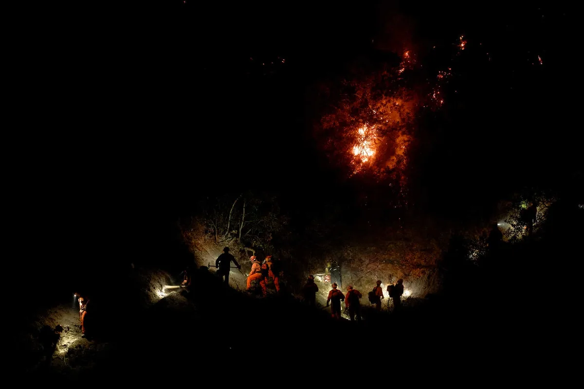 A nighttime photo shows silhouettes of firefighters in orange gear battling a raging wildfire, with flames illuminating the…