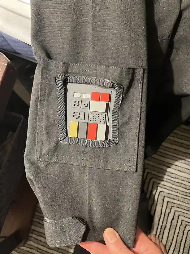 A close-up photo shows a person holding the sleeve of a dark gray jacket, which has a custom patch sewn onto the pocket. The…