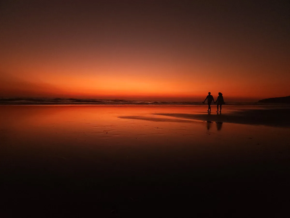 A romantic silhouette of two people holding hands and walking along a wet beach at sunset, with the sky and sand glowing in…