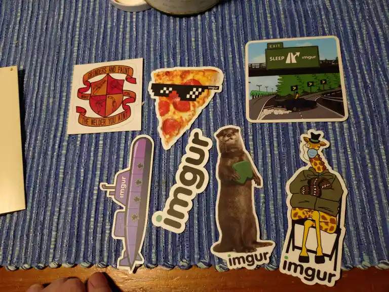 Several colorful stickers are arranged on a blue textured surface, featuring a pizza slice with pixelated sunglasses, a…