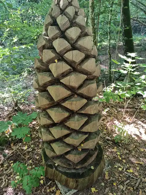 a tall, conical sculpture made of stacked, cut wooden segments stands in a forest, resembling a giant pinecone. The overlaid…