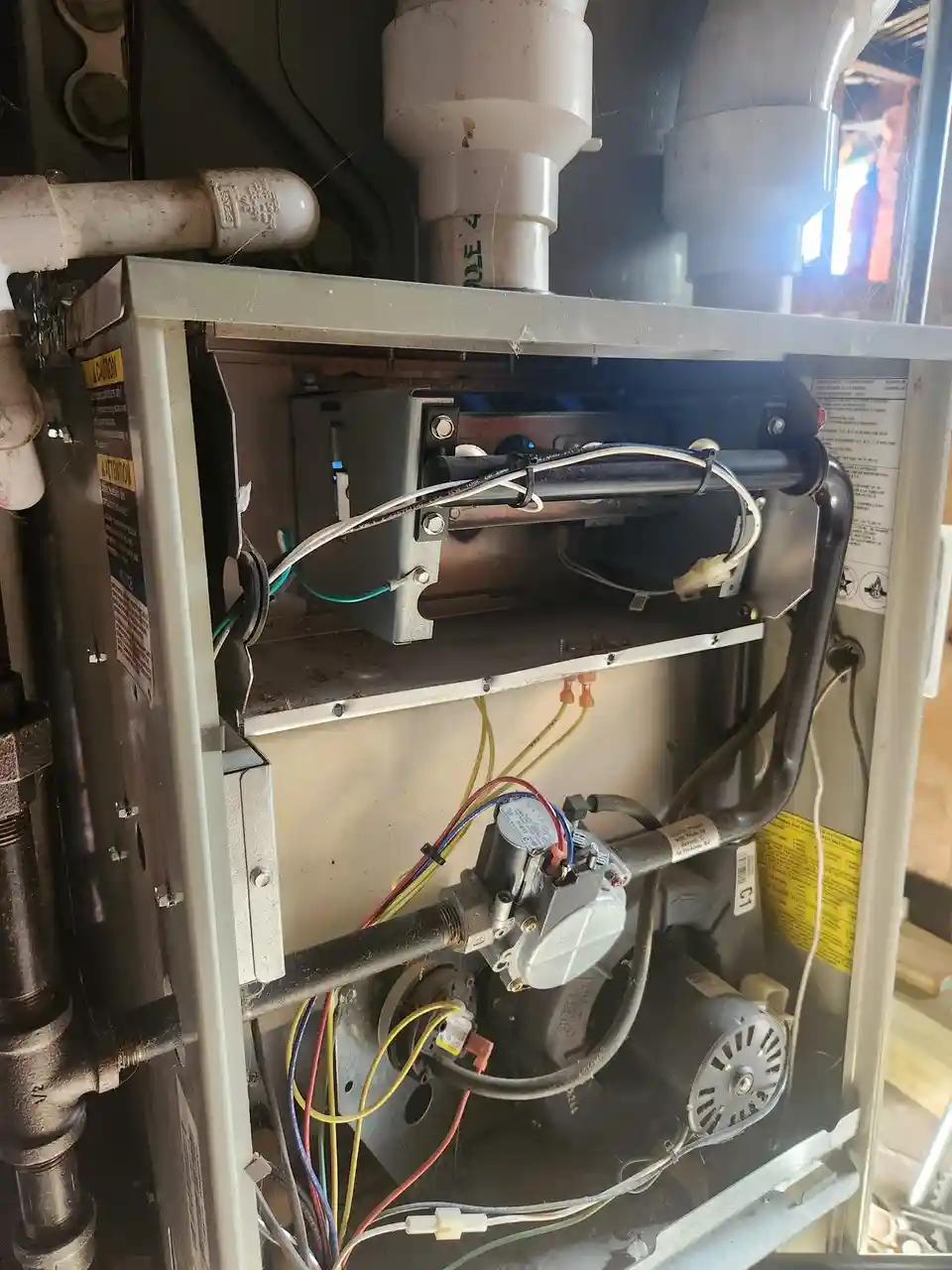 A close-up photo of the interior of a dirty, exposed furnace or HVAC unit with tangled wires, pipes, and mechanical parts…