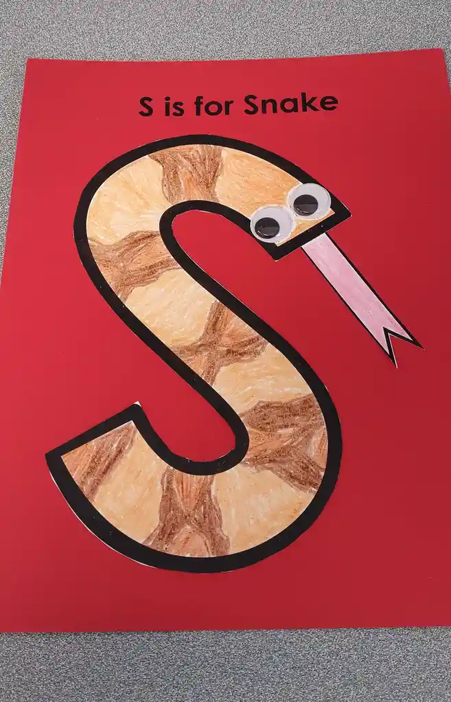 A child's craft project on red paper shows a large, black-outlined letter 'S' colored with brown crayon to look like a…