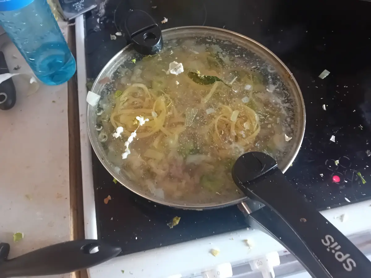 A messy, boiling pot of spaghetti sits on a black stovetop, with noodles, onions, and cheese floating in bubbling water…