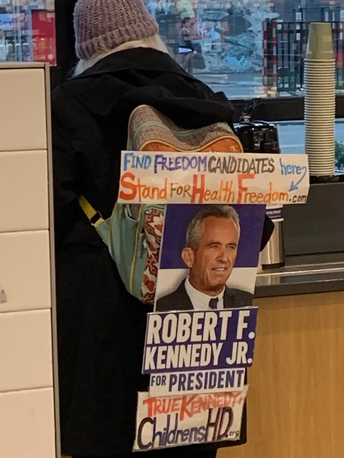 A person viewed from behind wears a backpack covered with multiple political and activist signs. The top sign reads 'FIND…