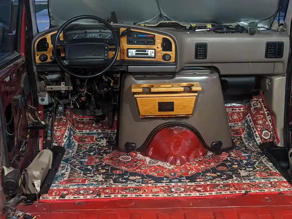 The interior of a stripped-down, red vehicle shows exposed wiring and a partially removed dashboard. The floor is covered…