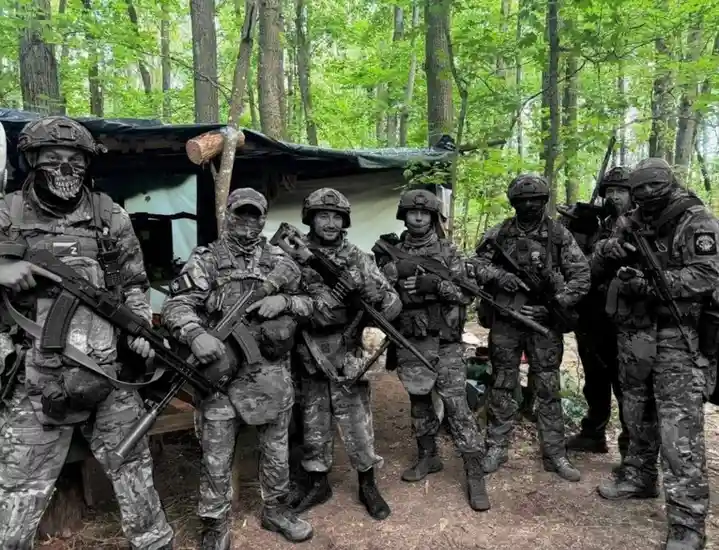 A group of seven men in full military camouflage gear, helmets, and face coverings, pose with rifles in a forest setting…