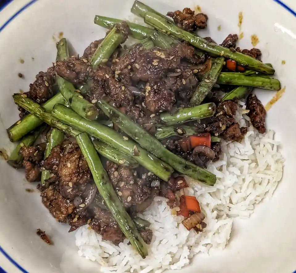 A close-up photo of a white bowl with a blue rim, containing a serving of white rice topped with stir-fried meat, green…