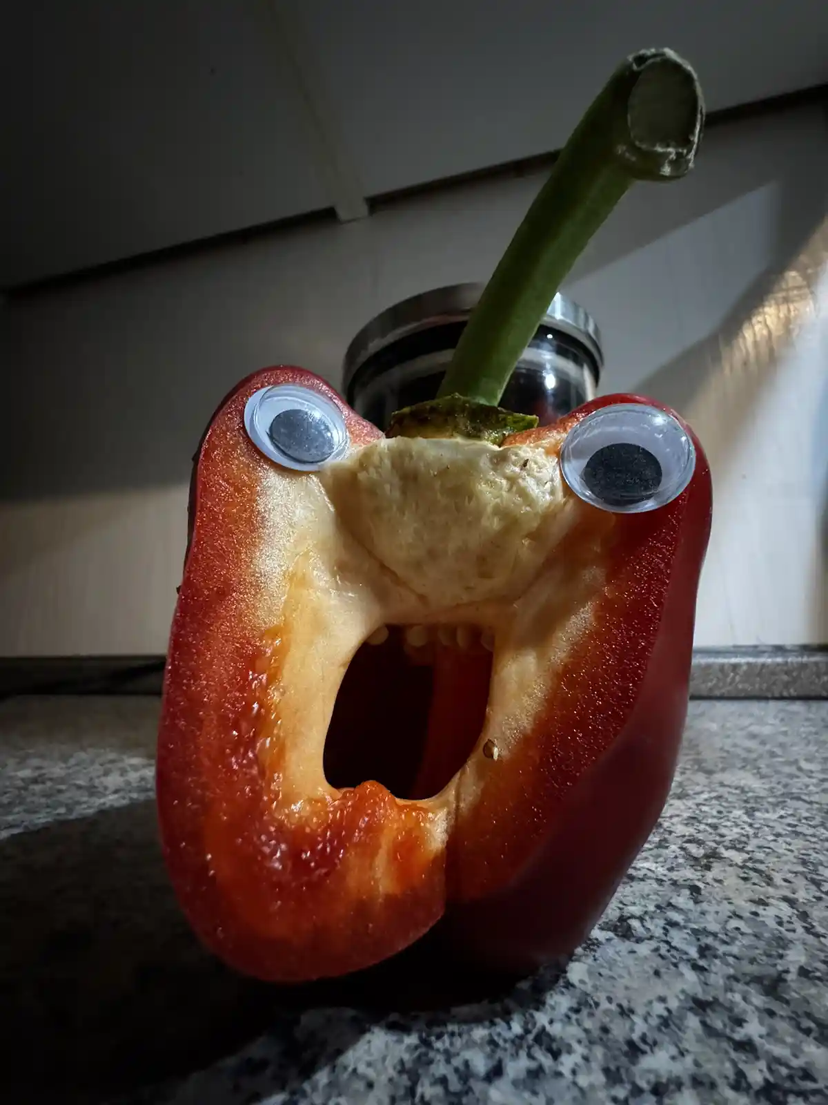 A red bell pepper, cut in half and standing upright on a countertop, has googly eyes glued to its cut edges, giving it a…