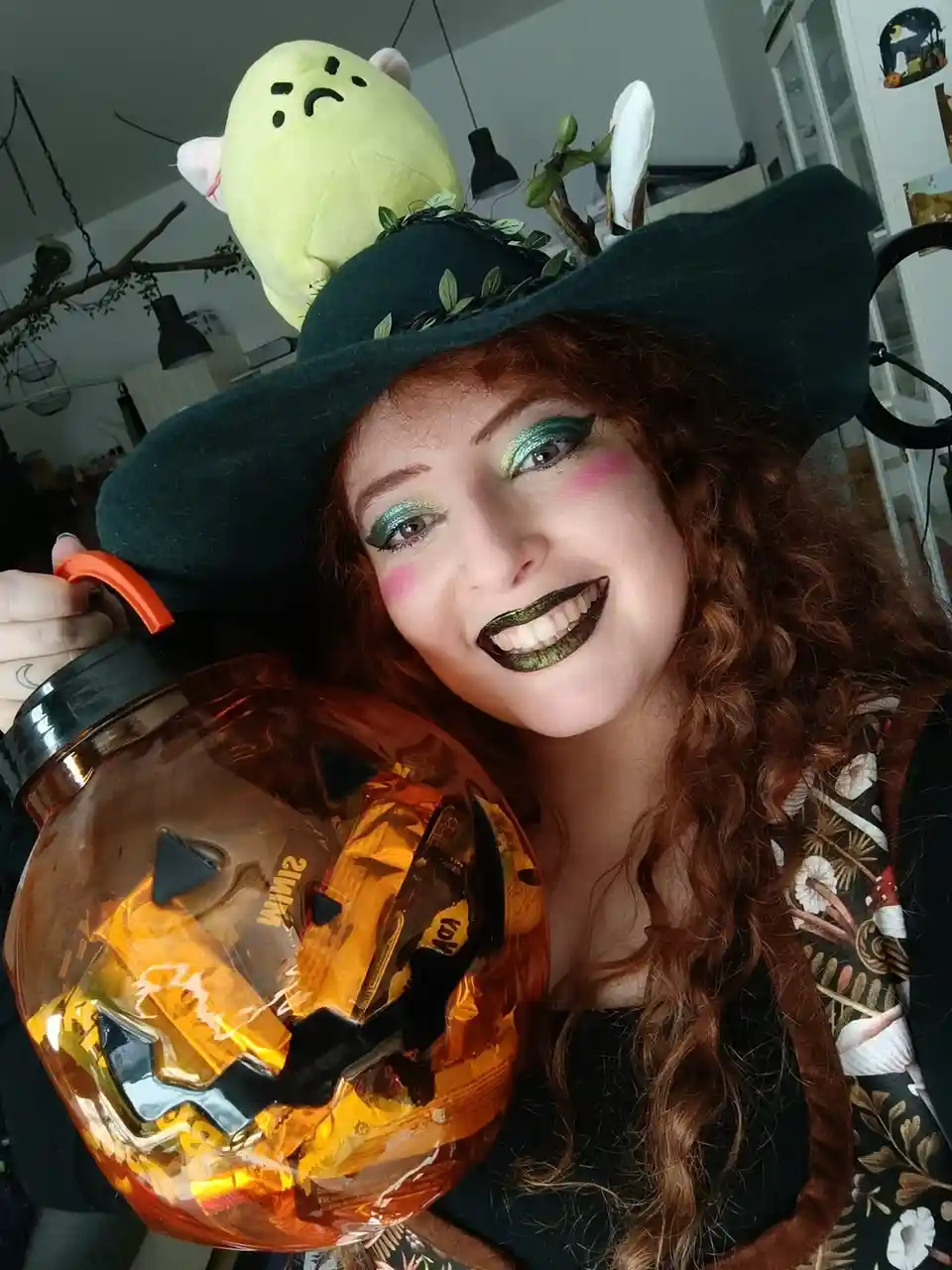A woman with curly red hair and witch-themed makeup, including green eyeshadow and dark glittery lips, smiles while holding…