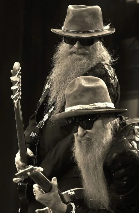 A black-and-white photo shows two identical-looking men with long beards, sunglasses, and fedoras, both holding electric…