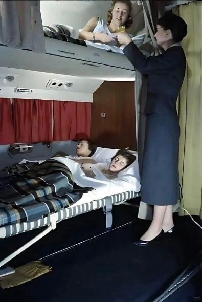 A vintage photo shows a flight attendant in a dark suit pouring a drink for a woman in a bunk bed on an old airplane. Below…