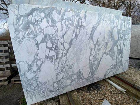 A large, rectangular slab of white and grey veined marble is propped up outdoors on wooden pallets, with trees and gravel…