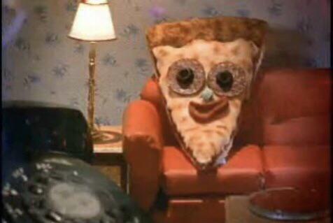 A slice of pizza with googly eyes and a smiling mouth sits in a red armchair in a dimly lit room with wallpaper and a lamp…