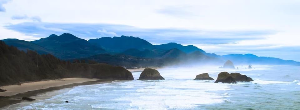 A scenic coastal landscape with waves crashing on a sandy beach, large sea stacks in the water, and distant mountains under…