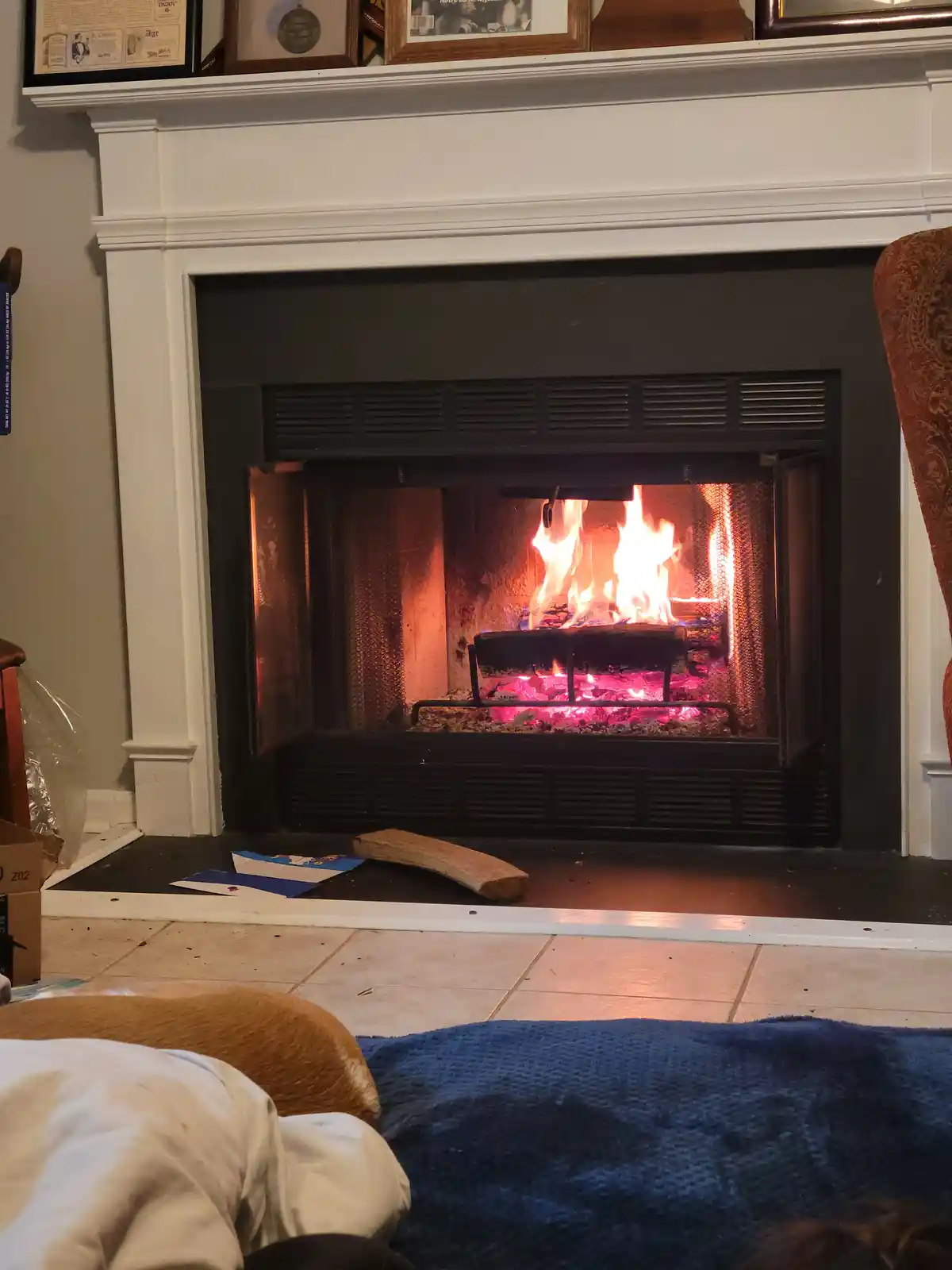 A cozy indoor scene shows a lit fireplace with flames and glowing embers inside a black metal firebox, framed by a white…