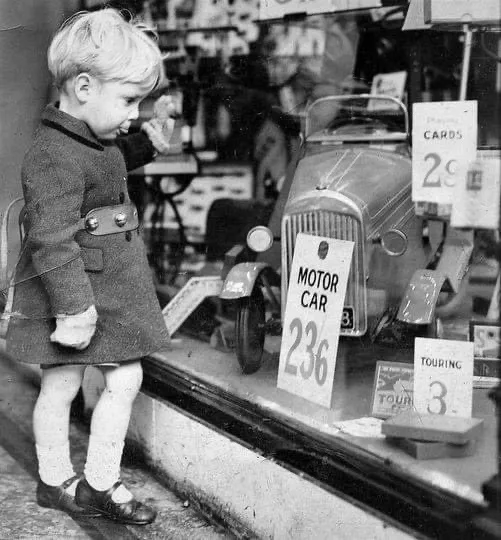 A young child in a vintage coat and white socks gazes intently at a toy motor car displayed in a shop window, priced at 236…