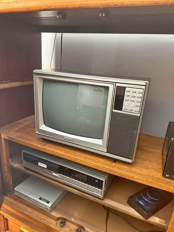An old silver CRT television sits on a wooden shelf, with a VCR and DVD player below it, and a modern black Apple AirPods…