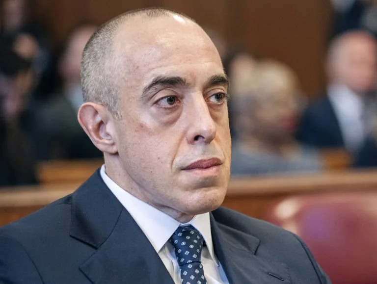 A close-up photo of a balding man in a dark suit and patterned tie, looking off to the side with a serious, slightly annoyed…