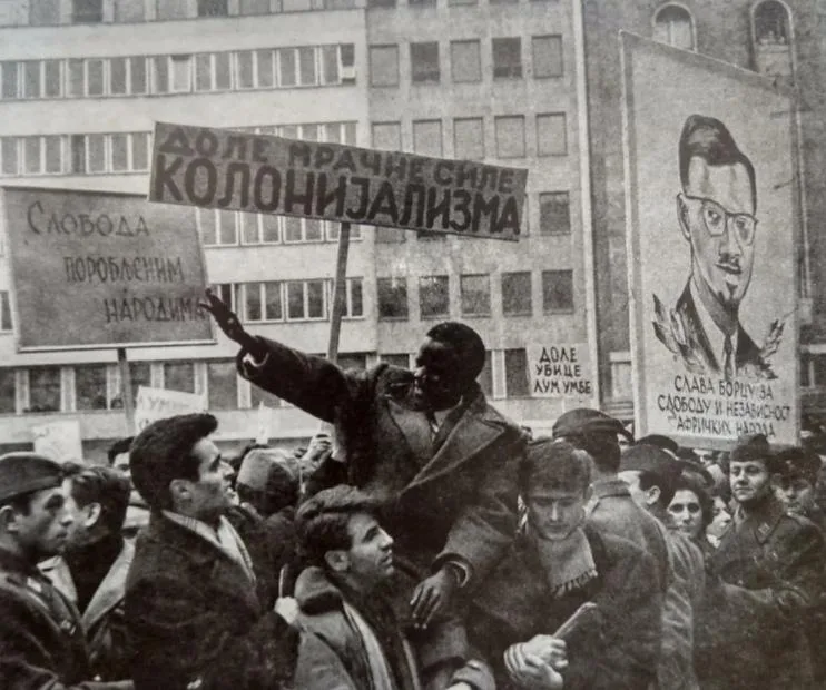 A black-and-white historical photo shows a Black man standing on shoulders in a crowd, holding a sign that reads 'ДОЛЕ…