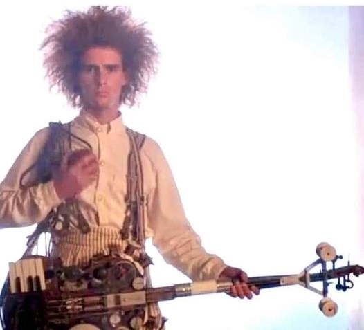 A man with wild, curly hair and a light-colored shirt stands against a bright background, holding a complex, steampunk-style…