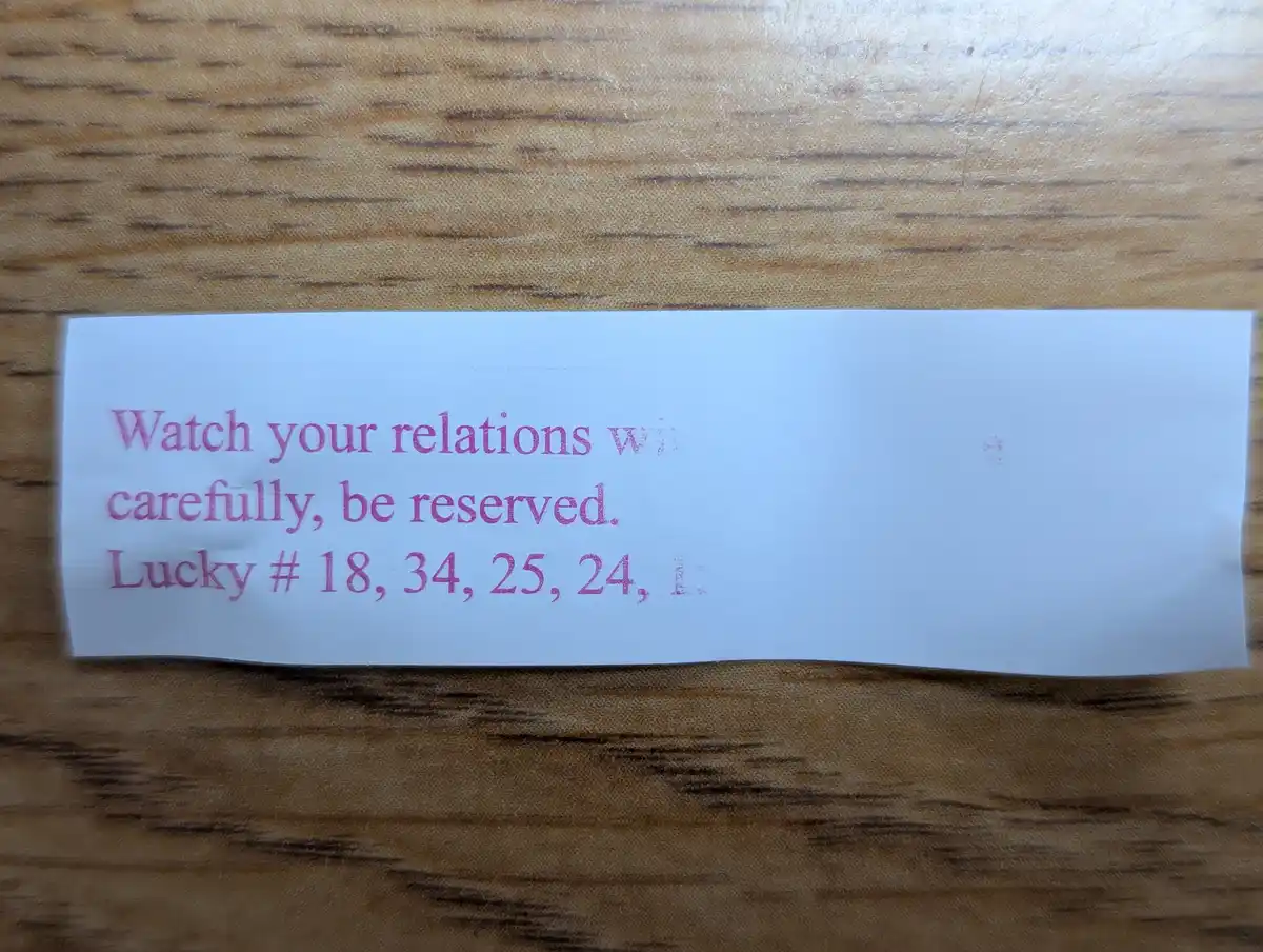 A white paper slip with pink text lies on a wooden surface, reading 'Watch your relations w carefully, be reserved. Lucky #…