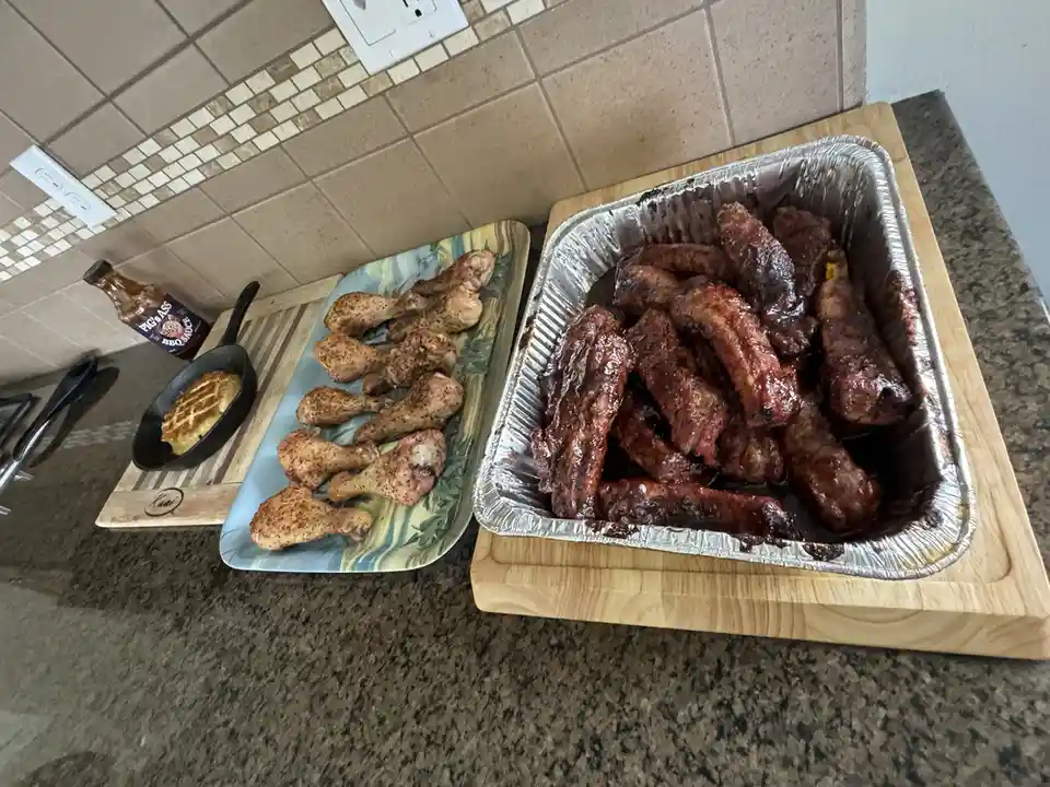 A kitchen counter displays a large aluminum tray filled with glossy, sauce-covered ribs and a rectangular plate with…