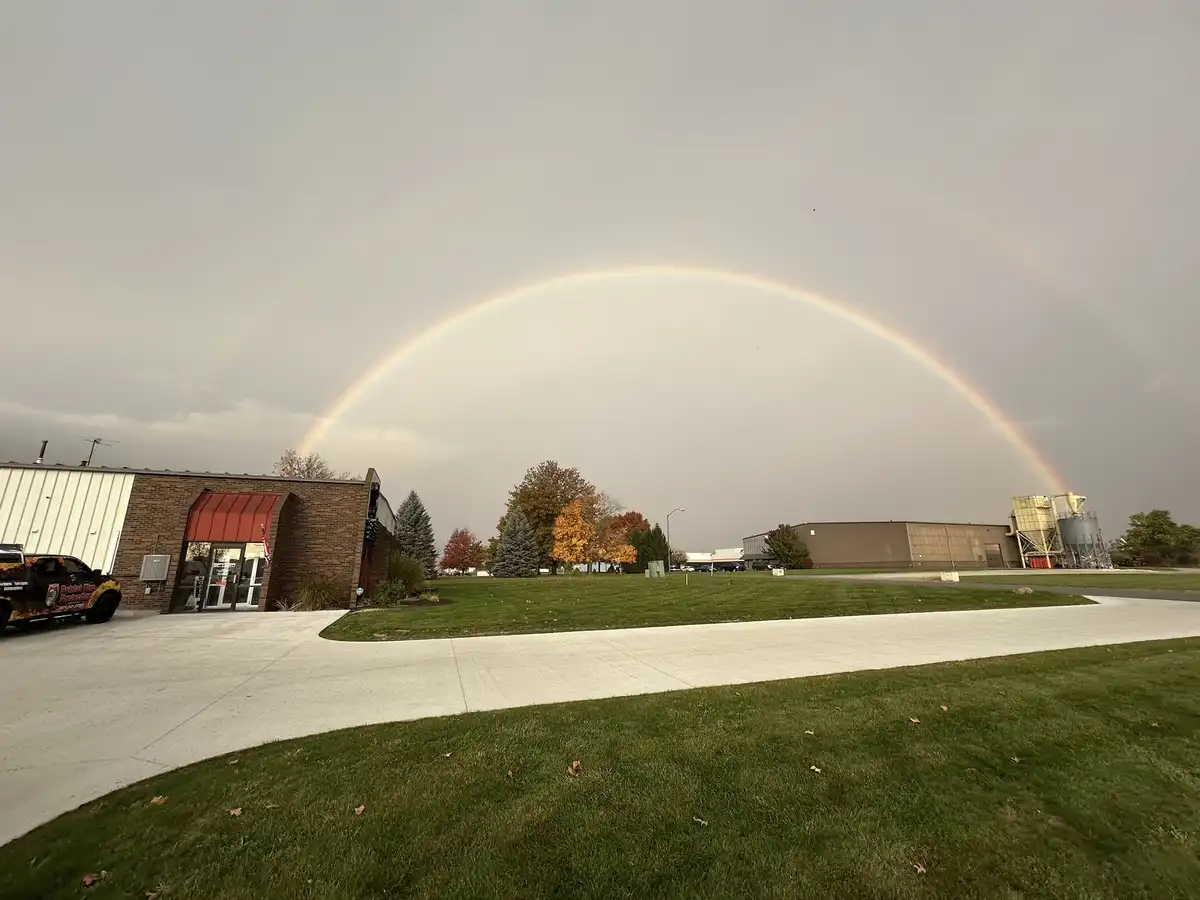 A wide-angle photo shows a vivid, full rainbow arcing across a gray, overcast sky above a grassy area with buildings and…