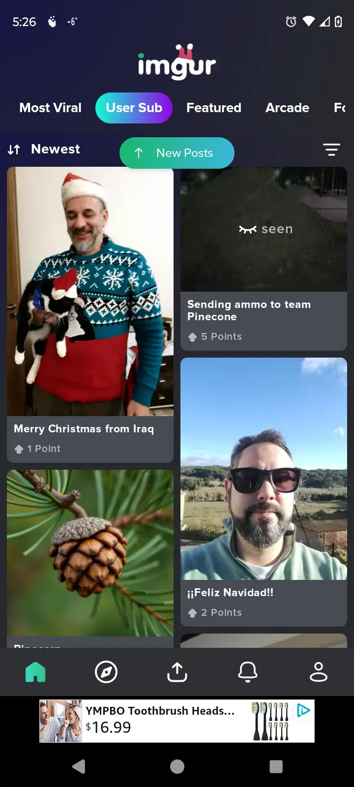 A man wearing a red and blue Christmas sweater and a Santa hat holds a black and white cat also wearing a tiny Santa hat…