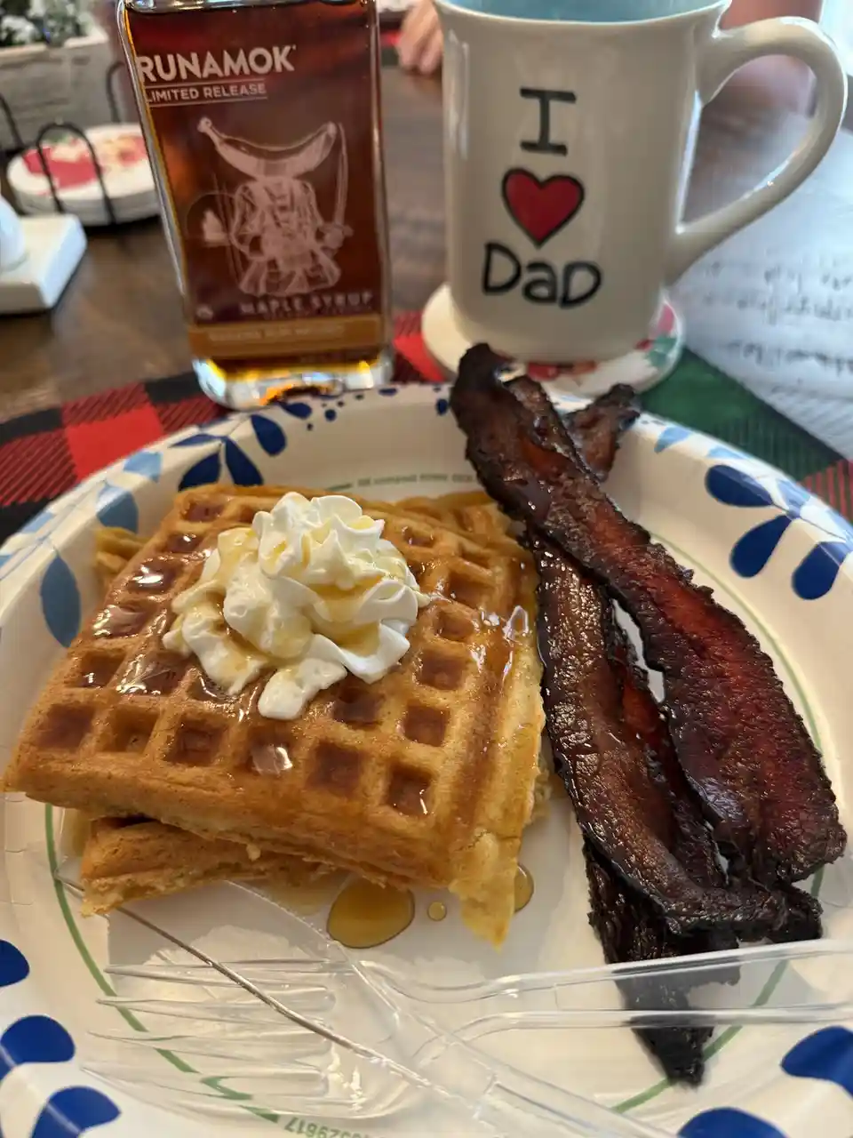 A close-up photo of a breakfast plate with waffles topped with whipped cream and syrup, alongside crispy bacon. Behind it…
