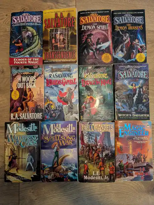 A collection of twelve fantasy paperback novels by authors R.A. Salvatore and L.E. Modesitt, Jr., arranged in three rows on…