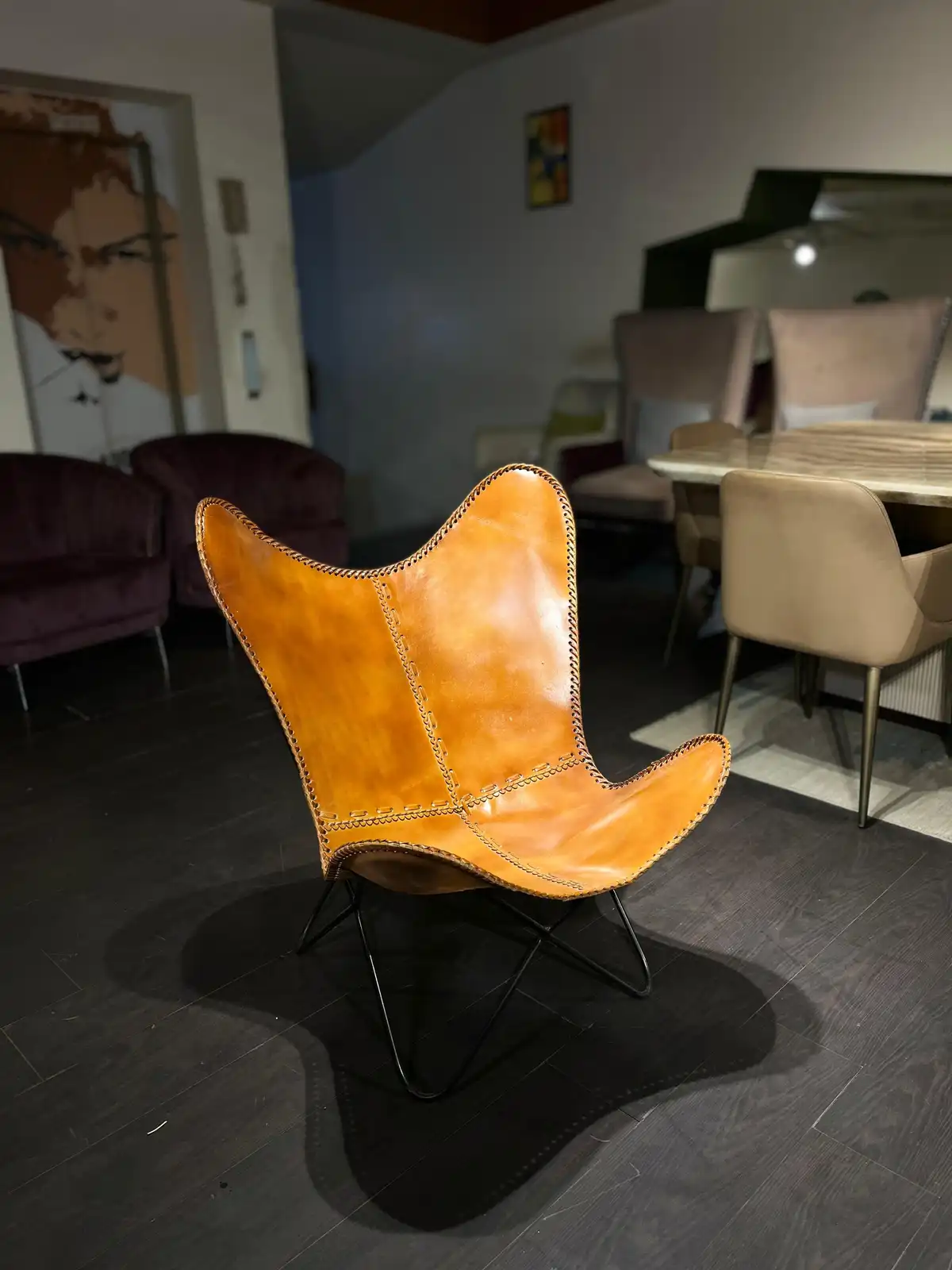 A tan leather butterfly chair with black metal legs sits in a modern furniture showroom on a dark wood floor, surrounded by…