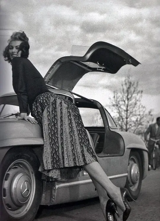 A vintage black-and-white photo shows a woman in a patterned skirt and dark top leaning against the open trunk of a classic…
