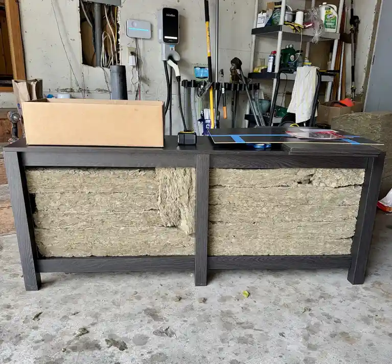 A dark gray modern desk sits in a cluttered garage, its front panels missing to reveal thick layers of beige fiberglass…