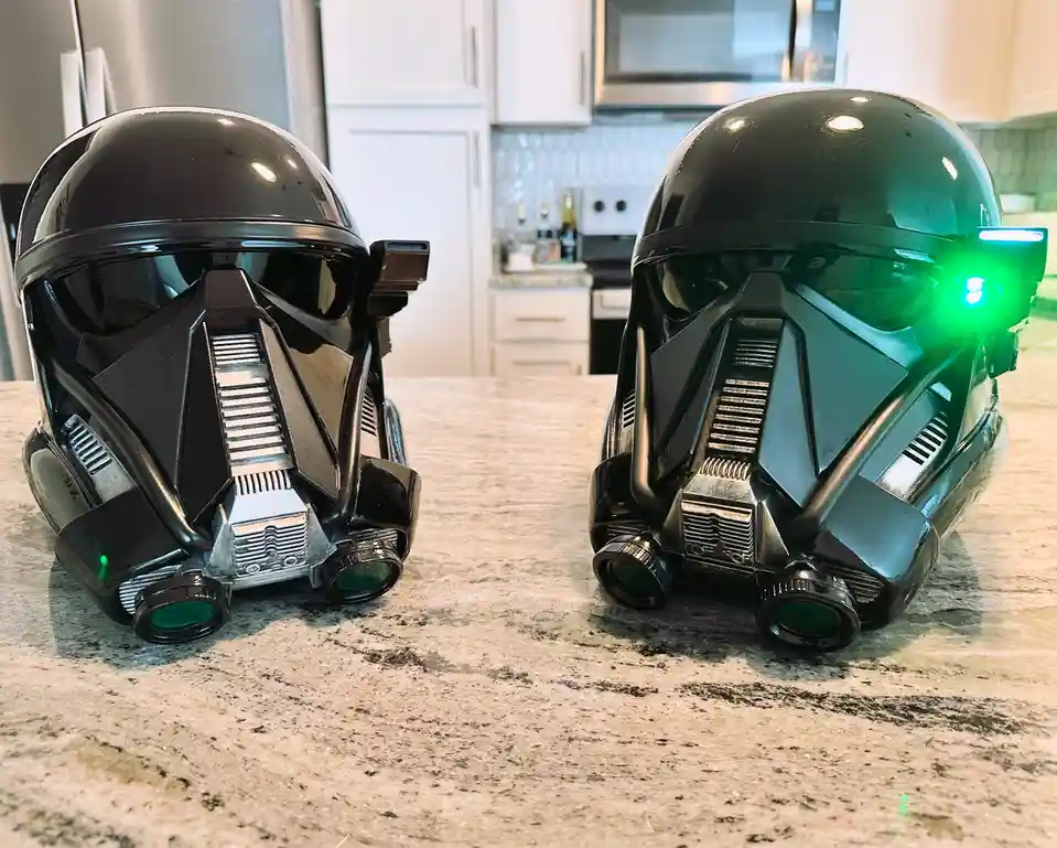 Two black Star Wars clone trooper helmets sit side-by-side on a kitchen countertop, one with glowing green lights on its…