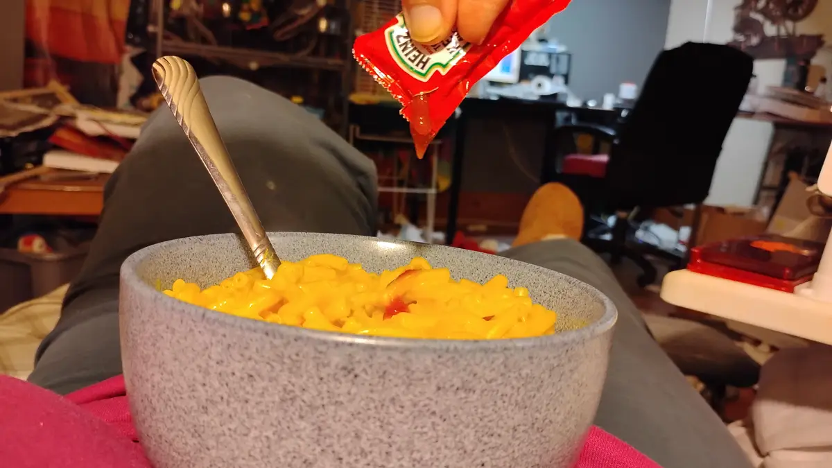 A person is pouring Heinz ketchup from a small red packet directly onto a bowl of macaroni and cheese resting on their lap…