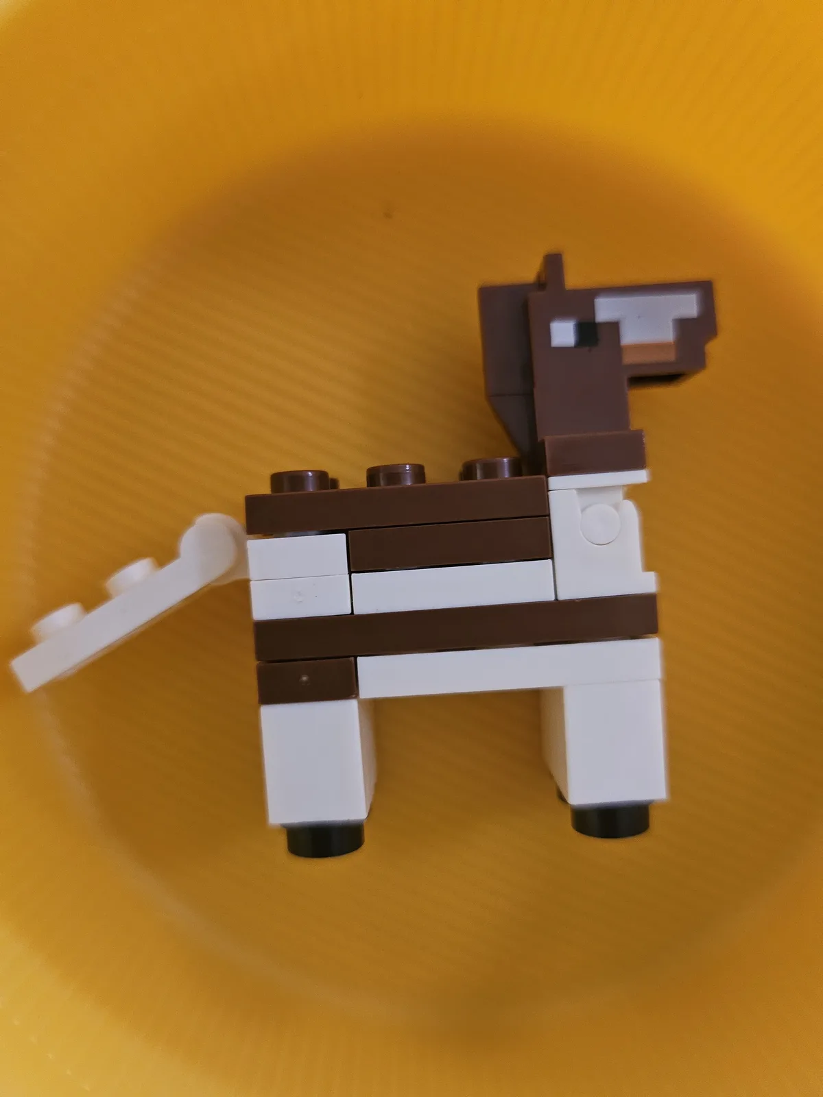 A handmade Lego figure resembling a llama, constructed from brown and white blocks with a pixelated head, stands on a…