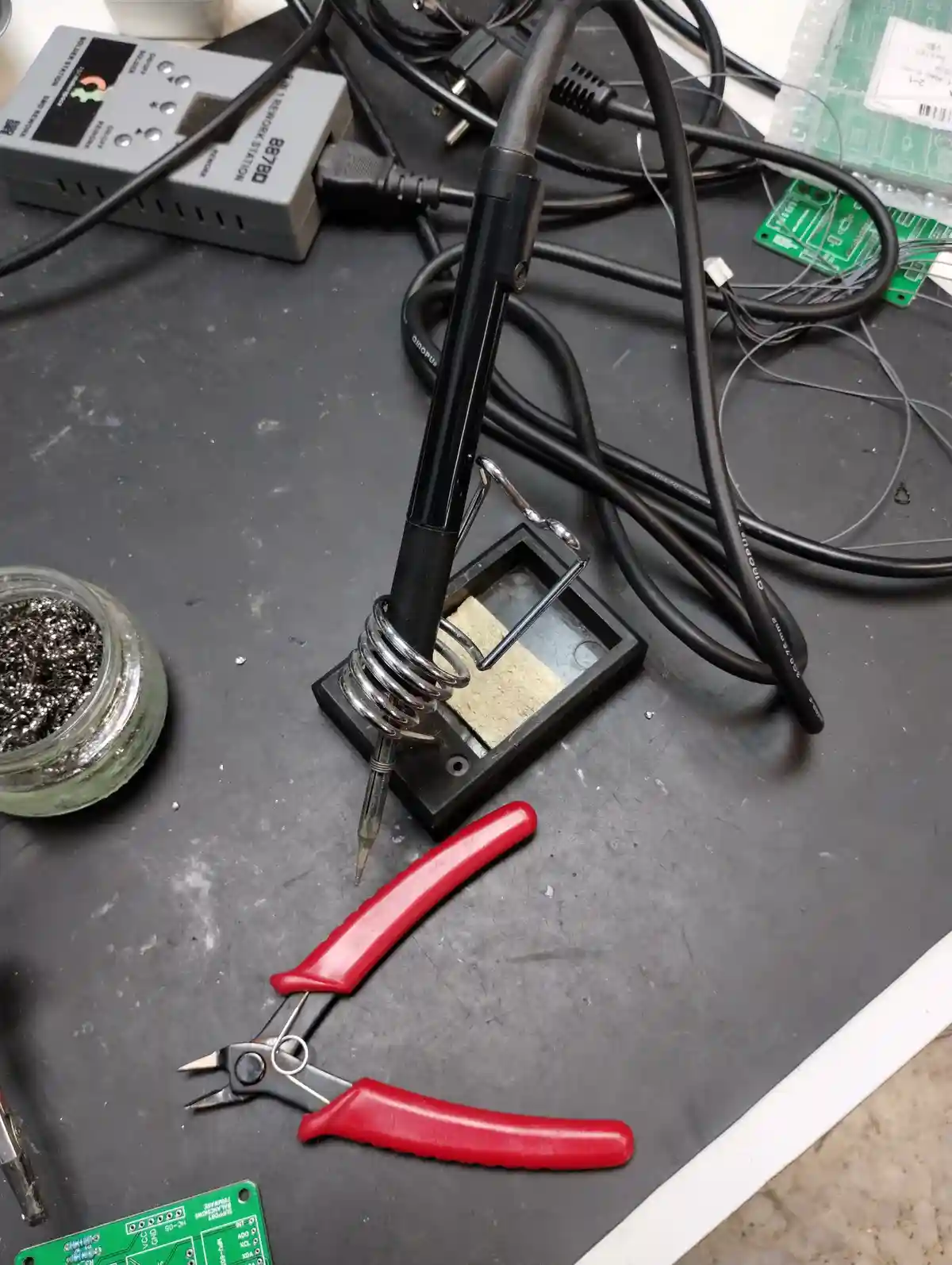A photo of an electronics workbench with a soldering iron in its stand, red-handled wire cutters, a jar of soldering flux…