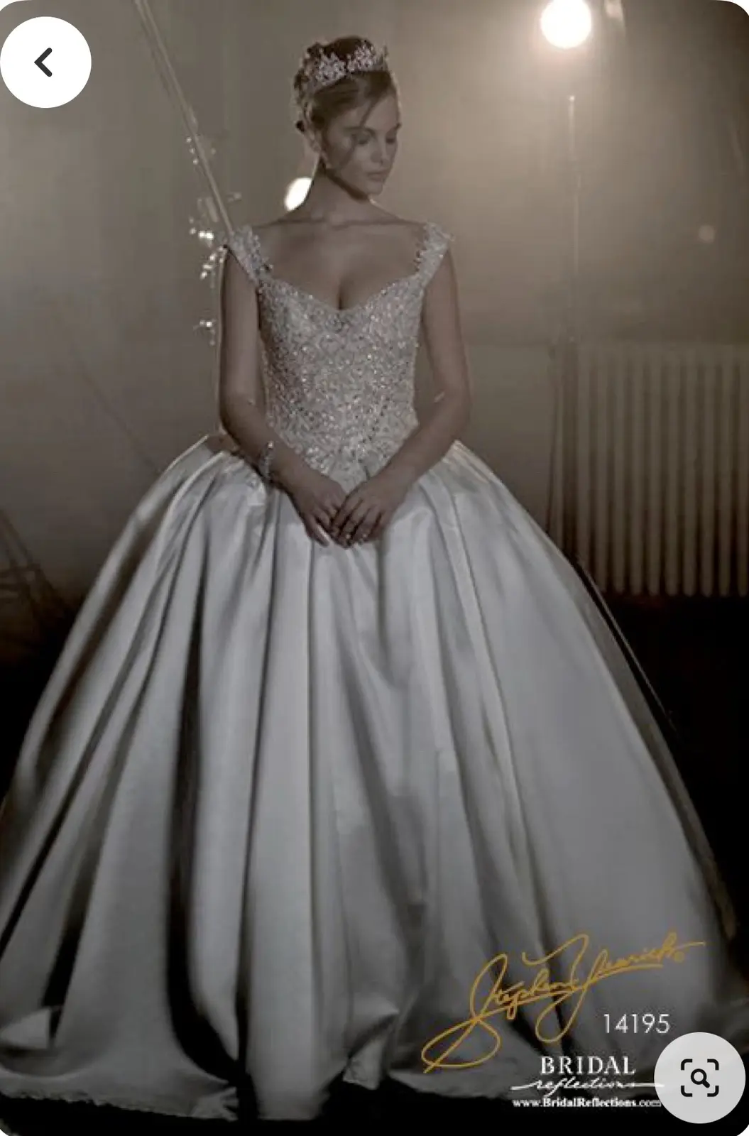A woman in a full-length, heavily embellished white wedding gown with a tiara, standing in a studio setting. The image has a…