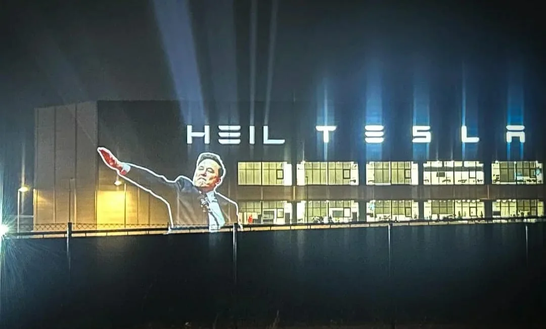 A nighttime photo of a large building with illuminated windows and spotlights. Projected onto the building's facade is a…