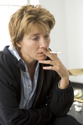 a woman with short brown hair, wearing a black zip-up jacket over a blue and white striped shirt, sits with a cigarette in…