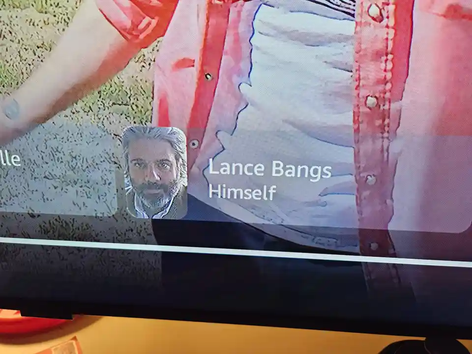 A close-up of a TV screen showing a person in a red shirt and white undershirt, with a small profile picture of a man with a…